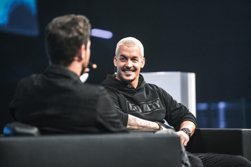 Dean is sitting on a stage in a black chair together with the show presenter. He is smiling towards the presenter. He is wearing a black hoodie from the brand Values Culture. In the hoodie the values LOYALTY is written in bold letters.